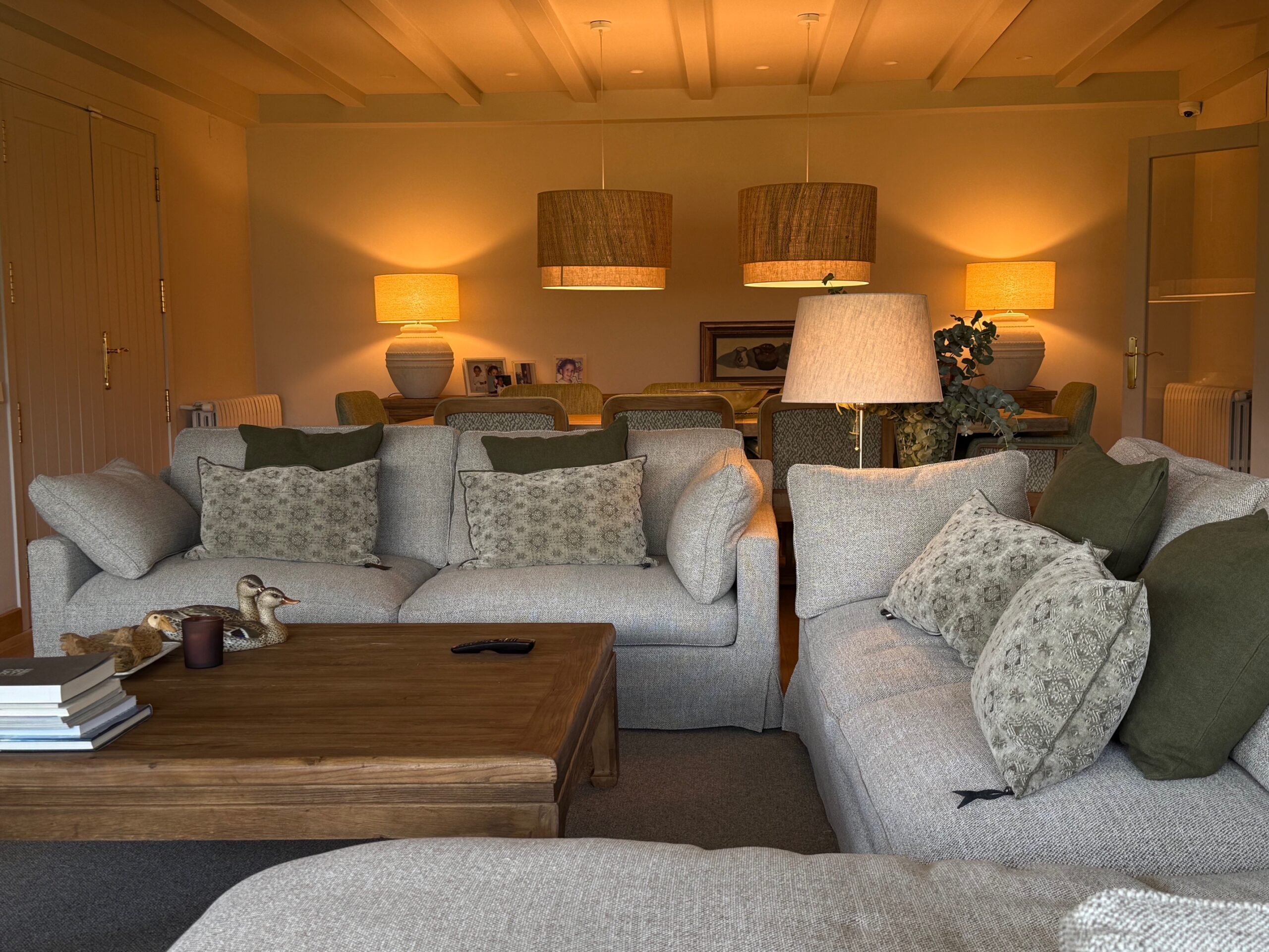 Sala d’estar acollidora amb sofàs en tons neutres, coixins decoratius, taula de centre de fusta i il·luminació càlida, oberta a un menjador amb llums penjants.