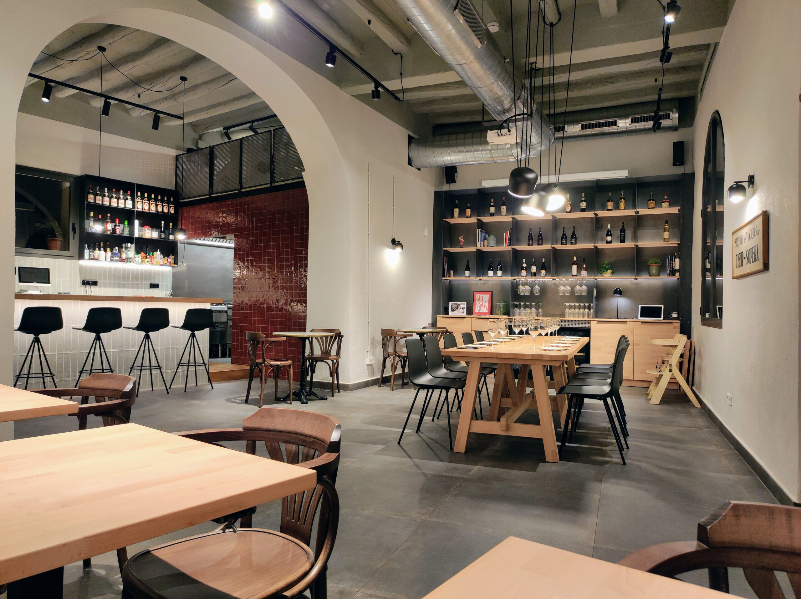 Interior de restaurant amb estil modern-industrial, amb barra amb tamborets, arc arquitectònic, taules de fusta i prestatgeries amb ampolles i copes, il·luminació càlida i conductes vistos al sostre.