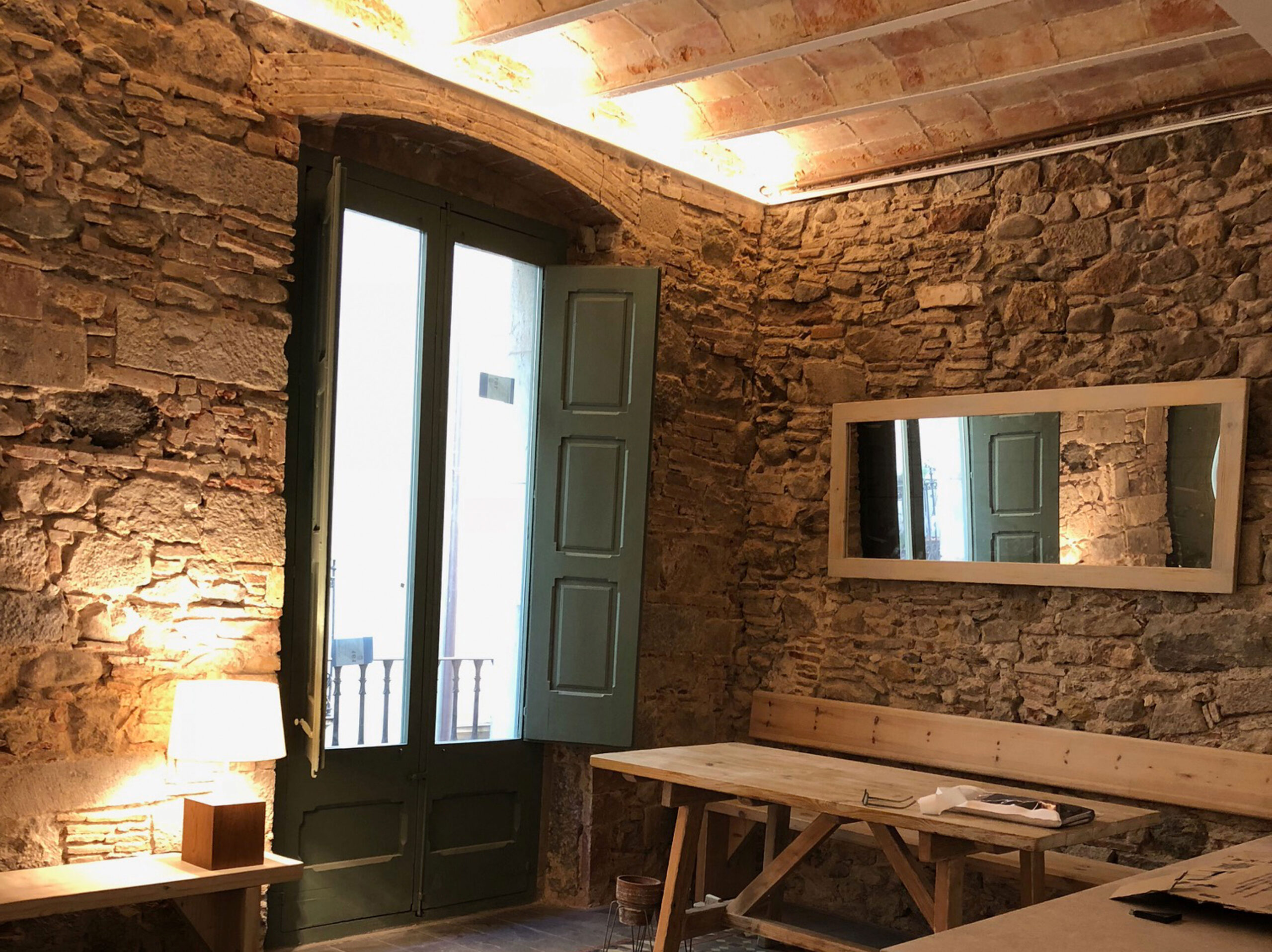 Interior amb parets de pedra vista i sostre amb volta, amb porta balconera verda, taula i banc de fusta, mirall i il·luminació càlida que ressalta l’estil rústic.