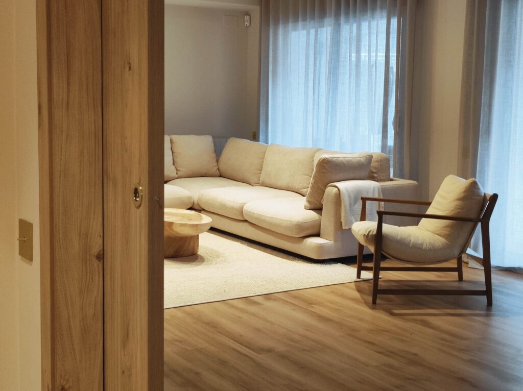 Sala d’estar càlida i minimalista amb sofà beix, butaca de fusta, taula de centre orgànica, cortines translúcides i terra de fusta natural.