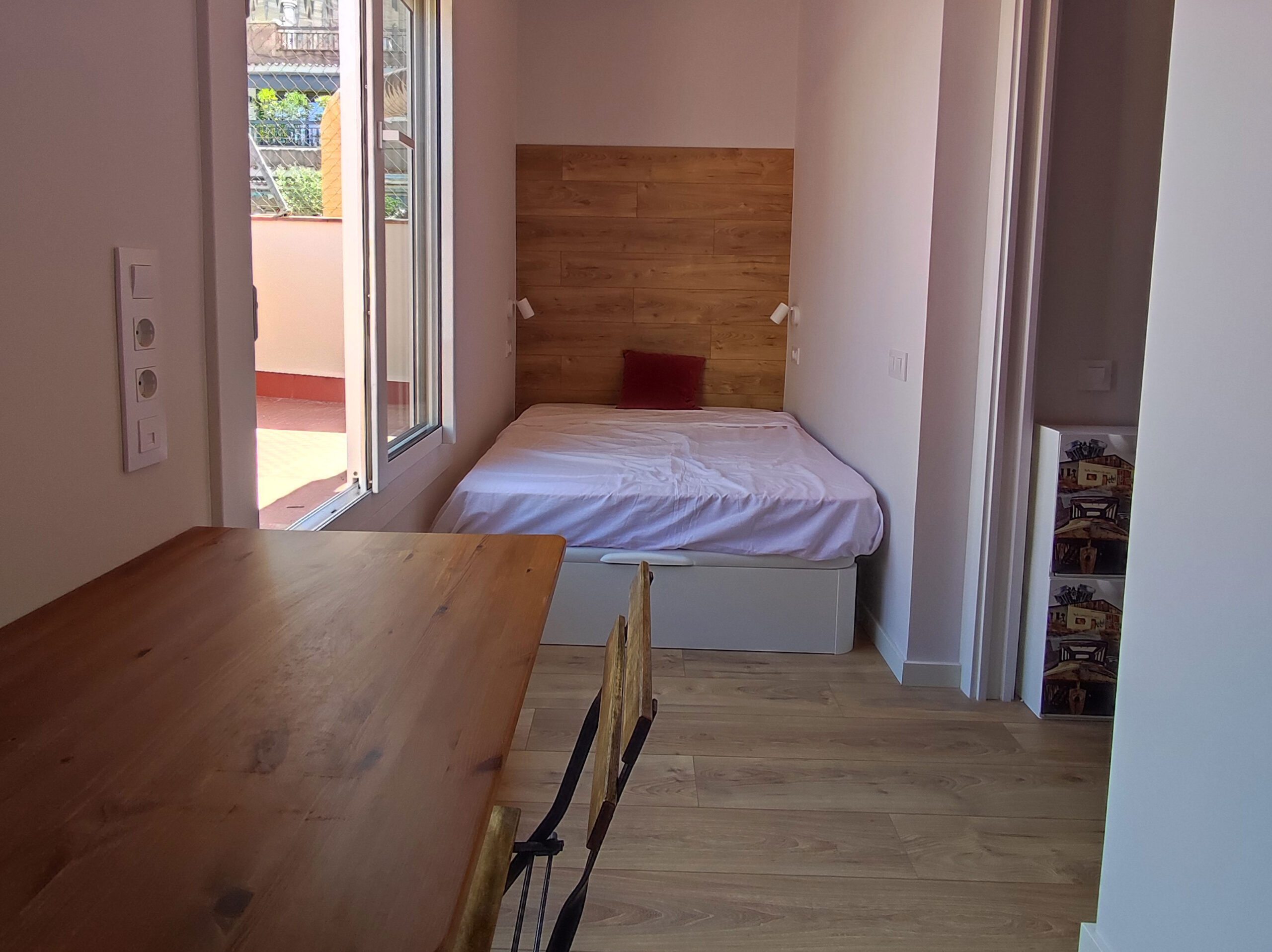 Habitació petita i funcional amb llit individual, capçal de fusta, escriptori compacte, terra de parquet i finestra amb sortida a balcó que aporta llum natural.